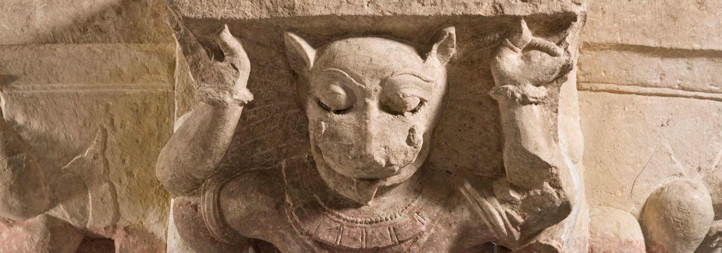 A pillar capital carved in the shape of a mythical being with an animal-like face, its hands appearing to hold a stone slab.