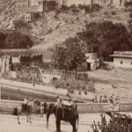 A black and white photograph of the Amber Fort, Jaipur, depicting bullock carts, camels and an elephant near a waterbody.