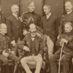 A black and white group photograph of sixteen men in two rows, with seven seated and nine standing behind them.