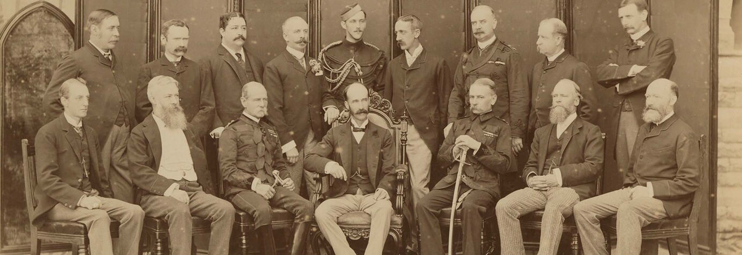 A black and white group photograph of sixteen men in two rows, with seven seated and nine standing behind them.