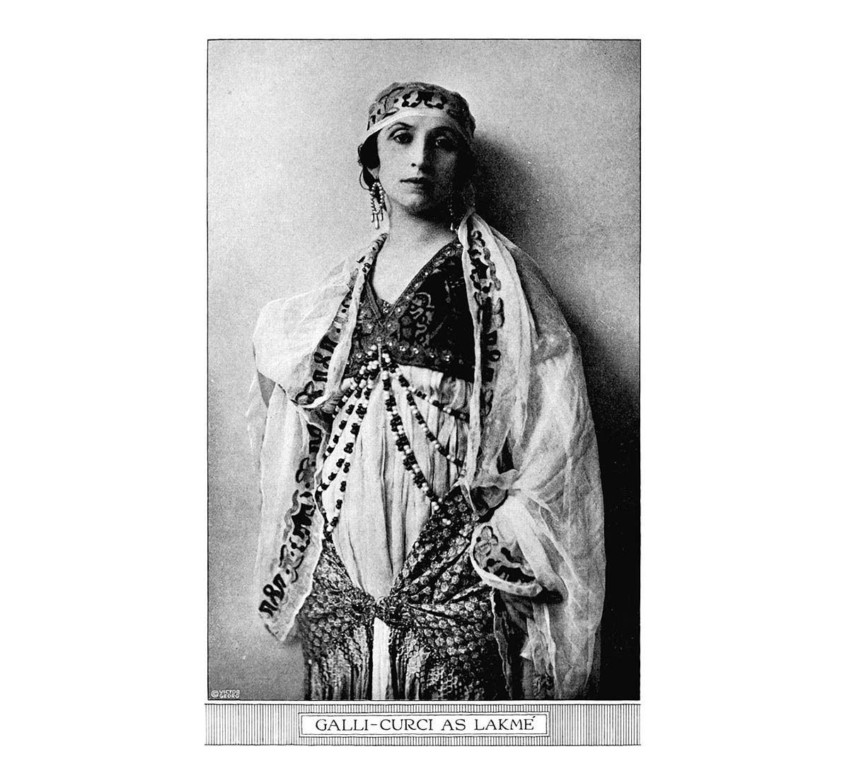 A photograph of a woman in ornamental headgear and outfit leaning against the wall.